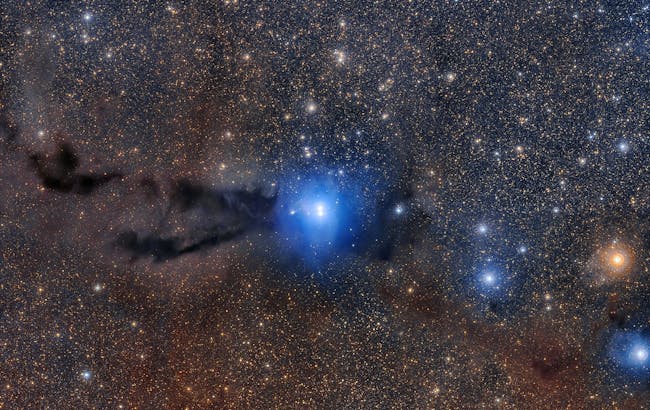 A dark cloud of cosmic dust snakes across this spectacular wide field image, illuminated by the brilliant light of new stars.