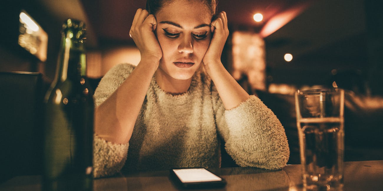 Depressed woman sitting in a bar and looking at her cell phone while waiting for a call from her boyfriend.